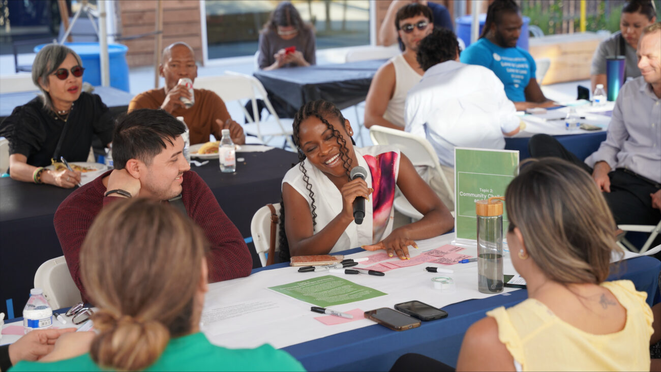 A group of people sitting at a table with a woman smiling with a microphone in her hand.
