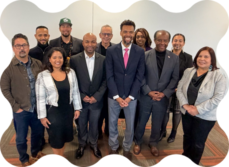 A formal group photo of professionals standing together in business attire. The group is arranged in two rows against a plain wall, showcasing a mix of suits and business-casual outfits.